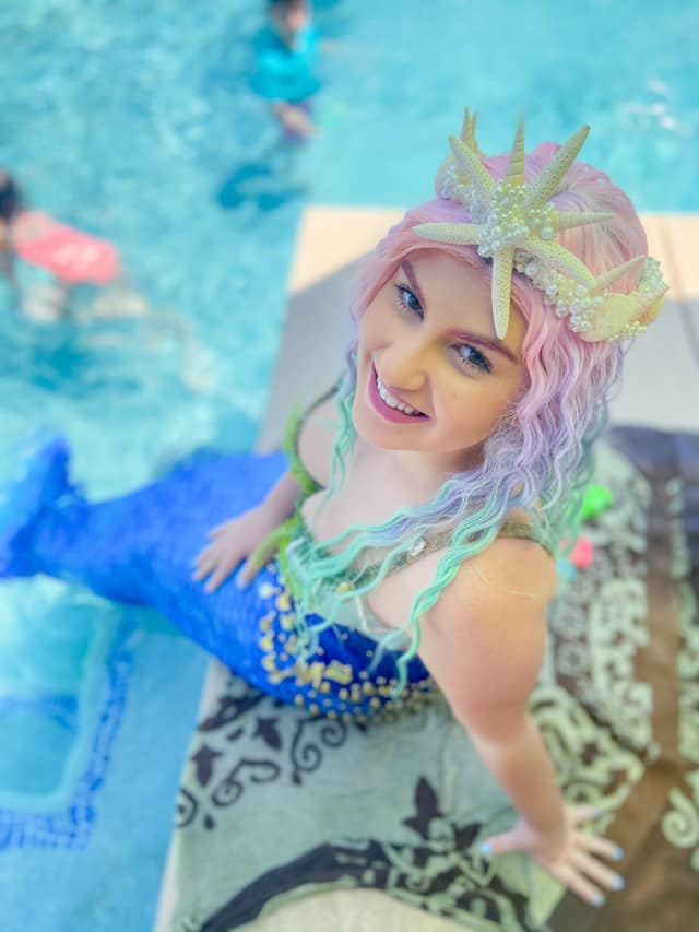 A pastel-haired mermaid performer with a starfish crown beside a pool